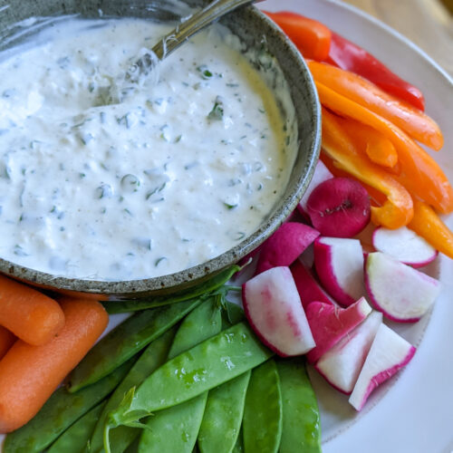 Parmesan Herb Veggie dip in a bowl surrounded by sliced bell pepper, radish, pea pods and carrots.