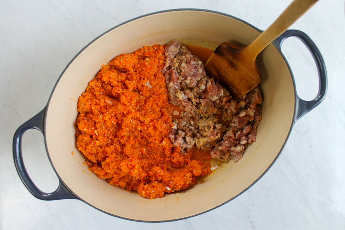 A large pot with raw Italian sausage cooking on one half and pureed vegetables sautéing on the other half.