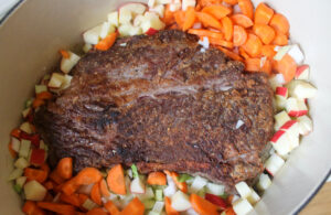 The beef roast searing in the pot surrounded by vegetables.