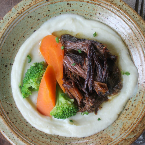 A plate of shredded beef roast on a bed of cauliflower puree with carrot and broccoli.