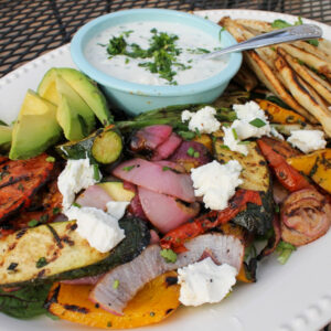 A white platter of grilled vegetables with pita and a bowl of yogurt sauce.