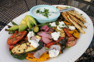 A grilled vegetables plate with crumbled goat cheese, avocado and a bowl of herb yogurt sauce.