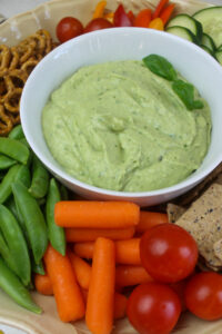 A bowl of avocado dip with carrots, pea pods and cherry tomatoes on a platter.