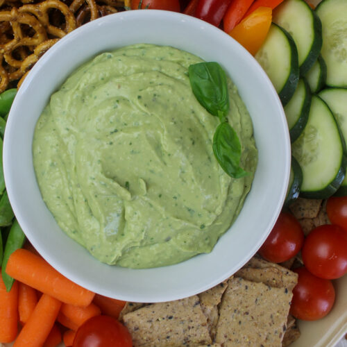 A bowl of green avocado bean dip with basil, surrounded by veggies, pretzels and crackers for snacking.