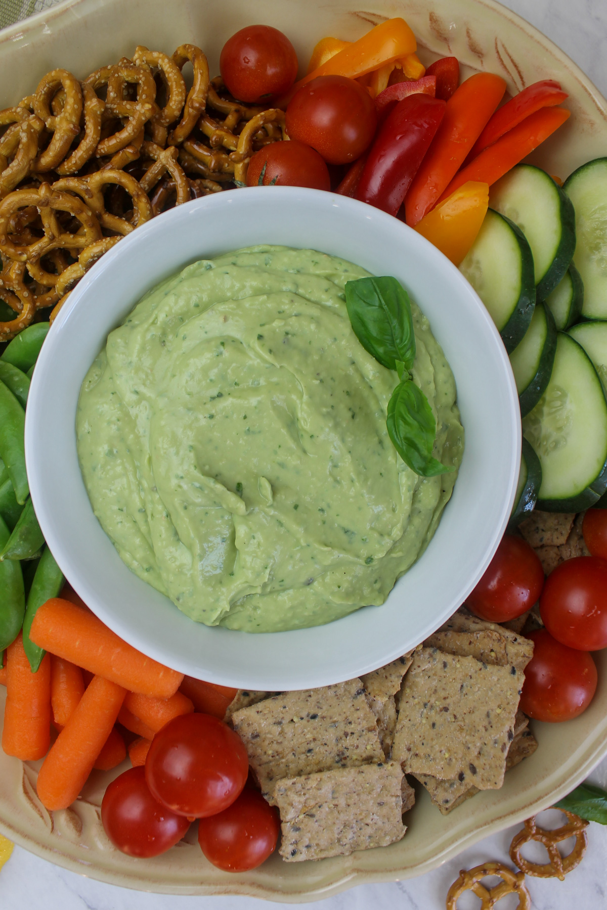 A bowl of avocado bean dip surrounded by a platter of raw vegetables with pretzels and crackers.