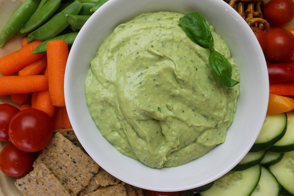 A close up of avocado basil dip with carrots, tomato, cucumber and pea pods around it.