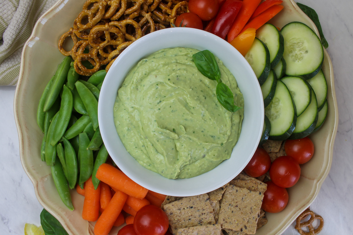 A white bowl of garlic avocado dip with sliced veggies and snacks.