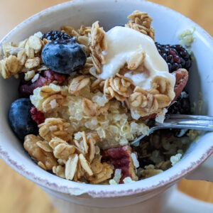 A white cup of breakfast quinoa porridge, with homemade granola, a scoop of yogurt and berries.