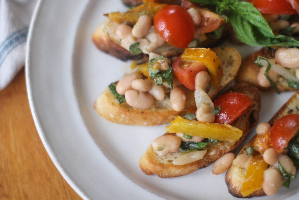 A white plate of white bean bruschetta on toasted crostini bread.