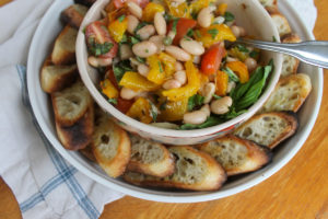 A bowl of white bean bruschetta with roasted pepper and tomato, with crostini around the plate.