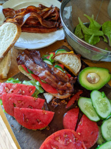 BLT's being prepared with extra veggies like tomato and cucumber slices, avocado and a plate of bacon.