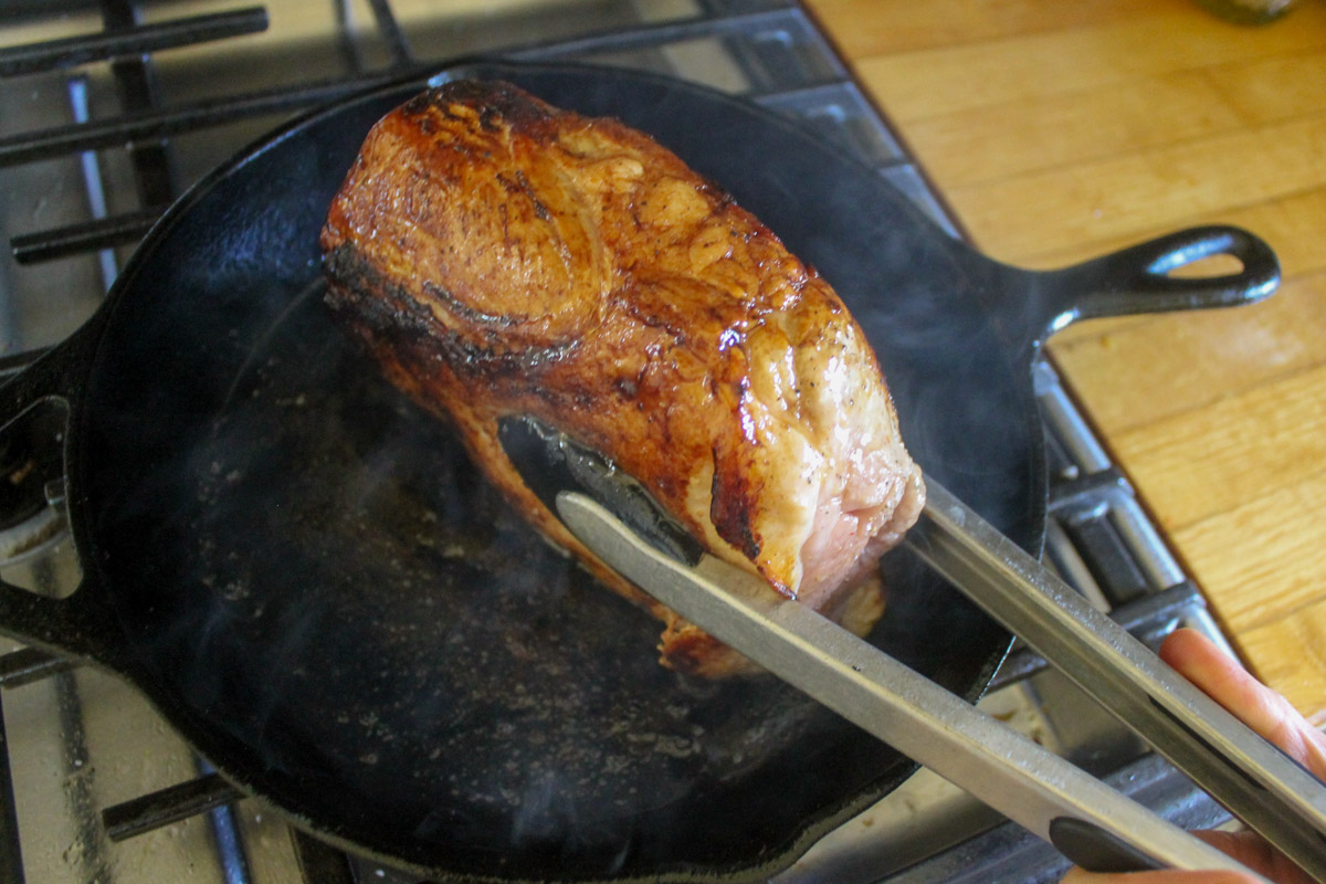 Searing a pork loin roast in a cast iron skillet, holding it up with tongs to sear the sides.