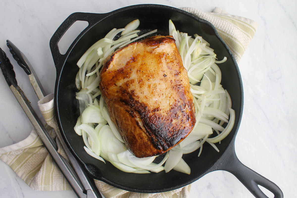 Adding raw sliced onions to the cast iron skillet surrounding the seared pork roast.