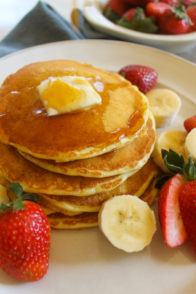 A stack of pancakes on a plate with butter, syrup, strawberries and banana.