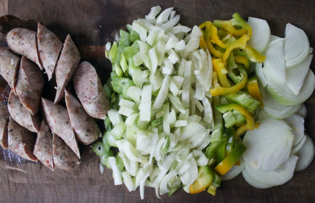 Ingredients, sliced onion, bell peppers, fennel and sausage links on a cutting board.