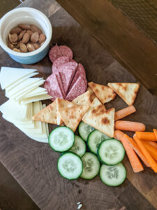 A simple lunch snack board with veggies, salami, cheese, pita crackers and a bowl of almonds.