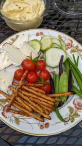 A charcuterie lunch plate with cherry tomato, cucumber, brie cheese, pretzels and pea pods with hummus.