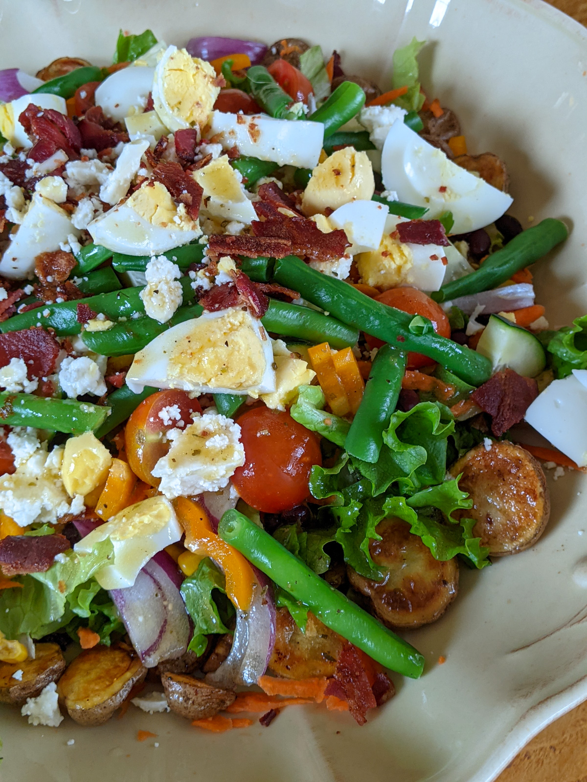 A platter of salad for dinner with green beans, hard boiled egg, bacon, on a bed of roasted potatoes.