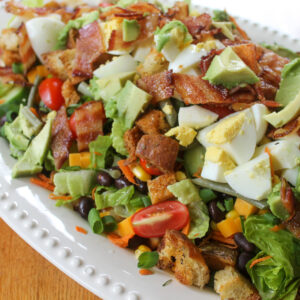 Green salad with potatoes, avocado, tomato, egg, bacon, and cheese on a dinner platter.