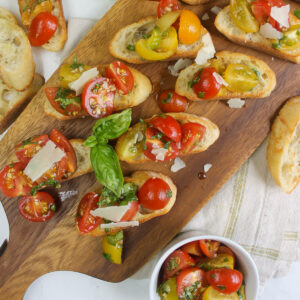 Bruschetta with cherry tomatoes on a wooden plank serving board with a bowl of extra topping.