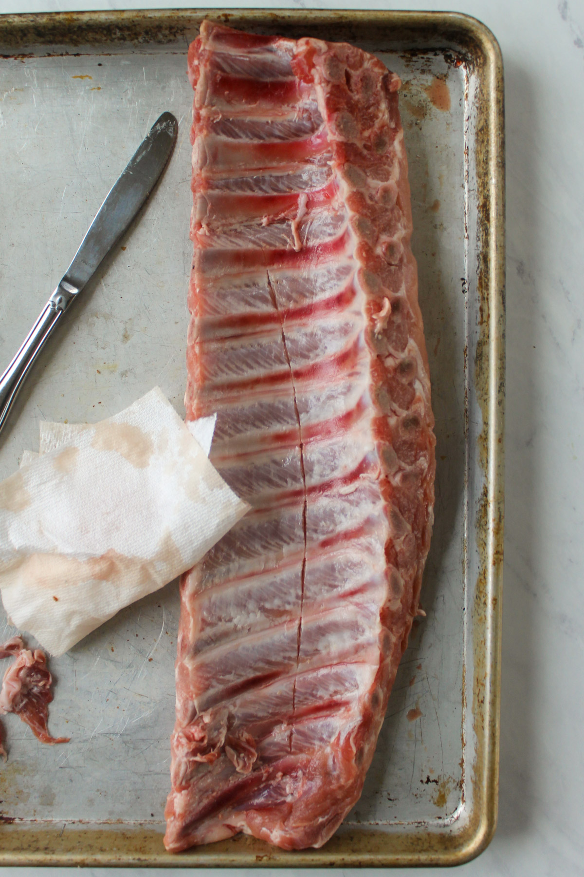 A rack of ribs on a sheet pan with a paper towel and a butter knife for removing the membrane.