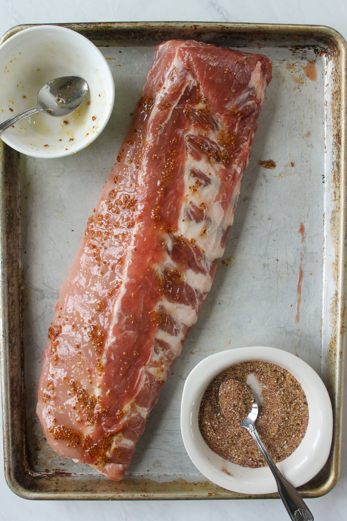 A rack of ribs on a sheet pan slathered with honey and mustard, by a bowl of dry rub.