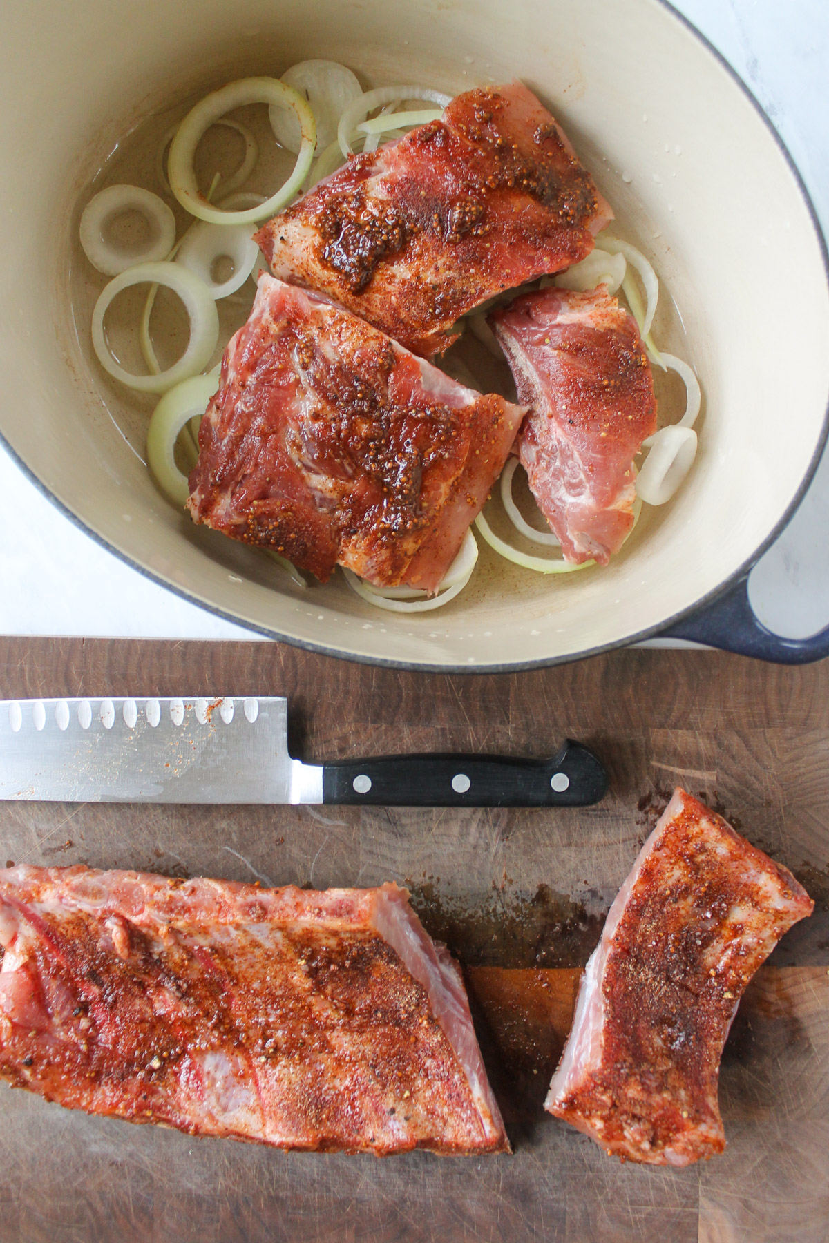 Cutting the dry rub ribs into portions on a cutting board and adding them to a Dutch oven on a bed of onions.