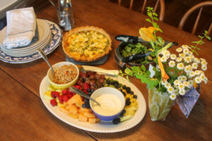 A table set for brunch with fruit, quiche, and salad, with a vase of flowers.