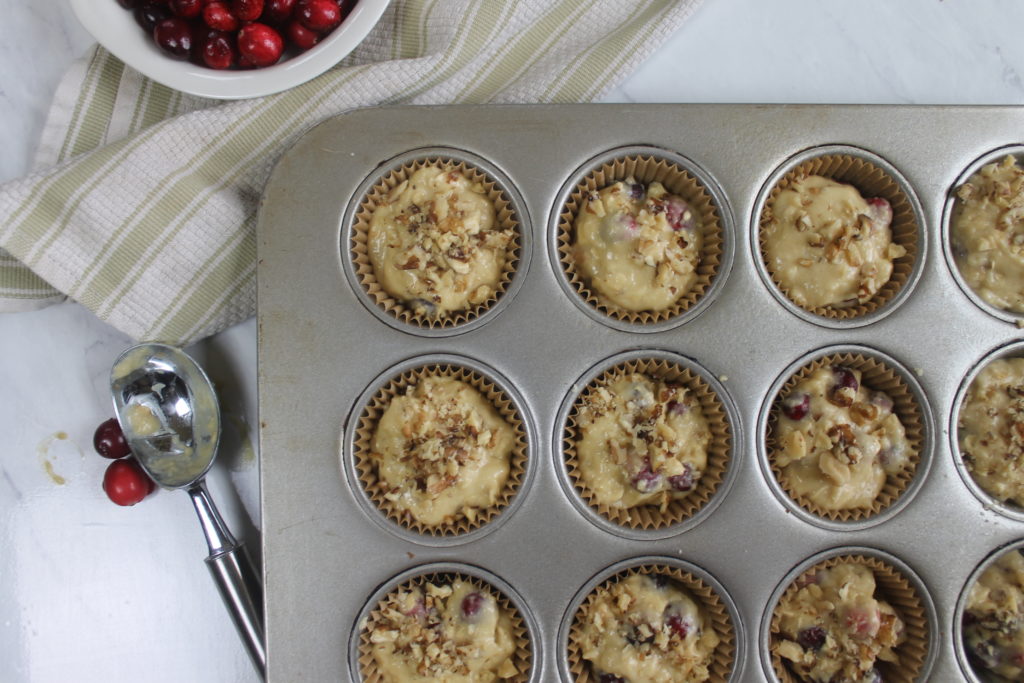 Cranberry Muffin batter in a lined muffin tin with a bowl of fresh cranberries and portion scoop. 