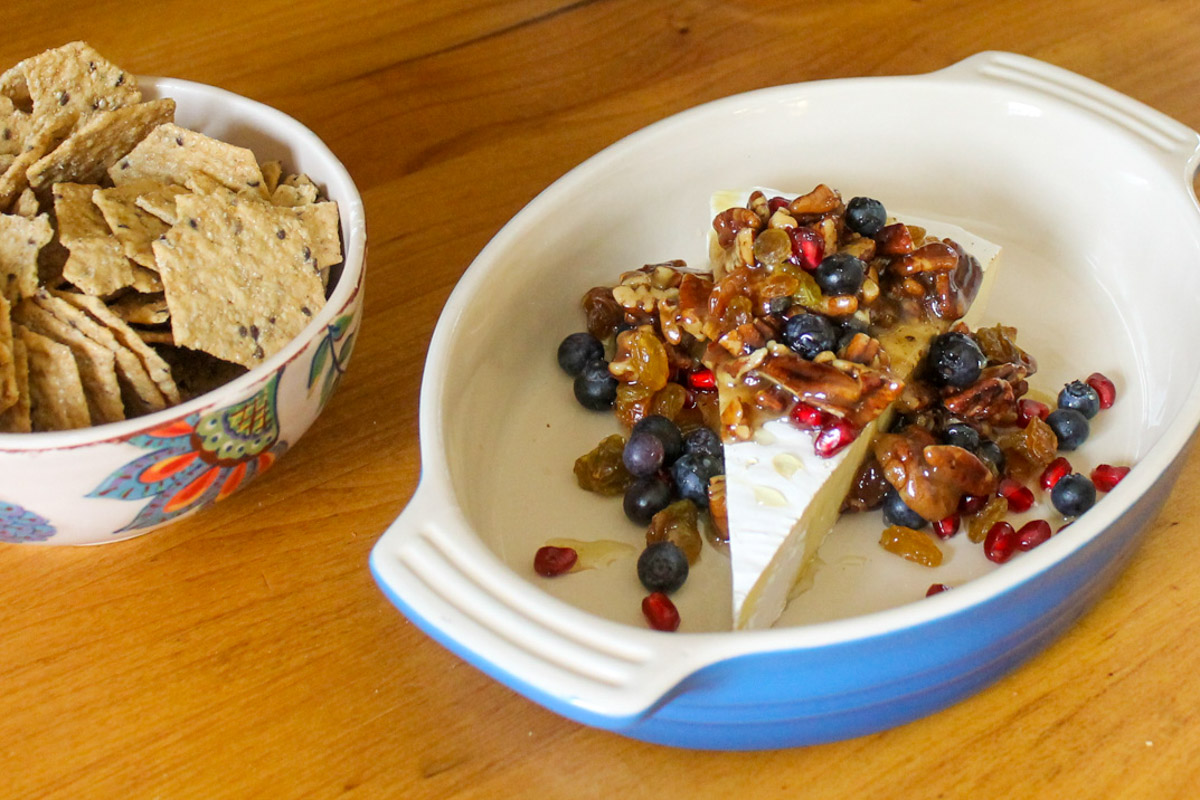 A blue oval dish with a wedge of brie topped with honey fruit and nuts with a bowl of crackers next to it.