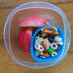 A cup of snacks with apple slices and a silicone muffin cup of homemade trail mix.