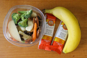 A to go snack cup of fruit and veggies next to 2 granola bars and a banana.