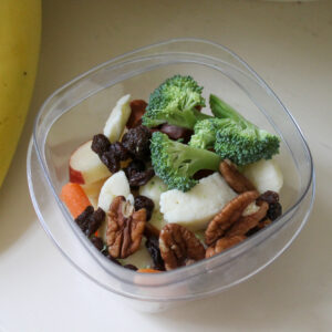 A cup of healthy kid snacks with broccoli, apple, cheese, raisins and pecans.