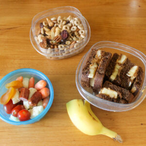 Healthy snacks for kids like trail mix, peanut butter banana sandwich and a cup of veggies with cheese curds and beef sticks. And half a banana.