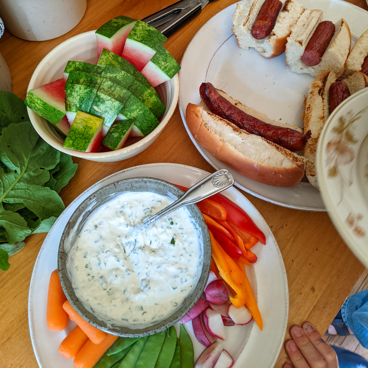 A kid friendly dinner on the counter with raw veggies and dip, hot dogs and watermelon slices.