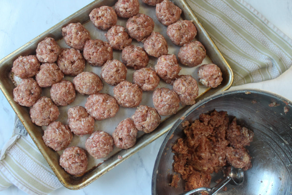 Raw meatballs on a sheet pan next to a bowl with a portion scoop.