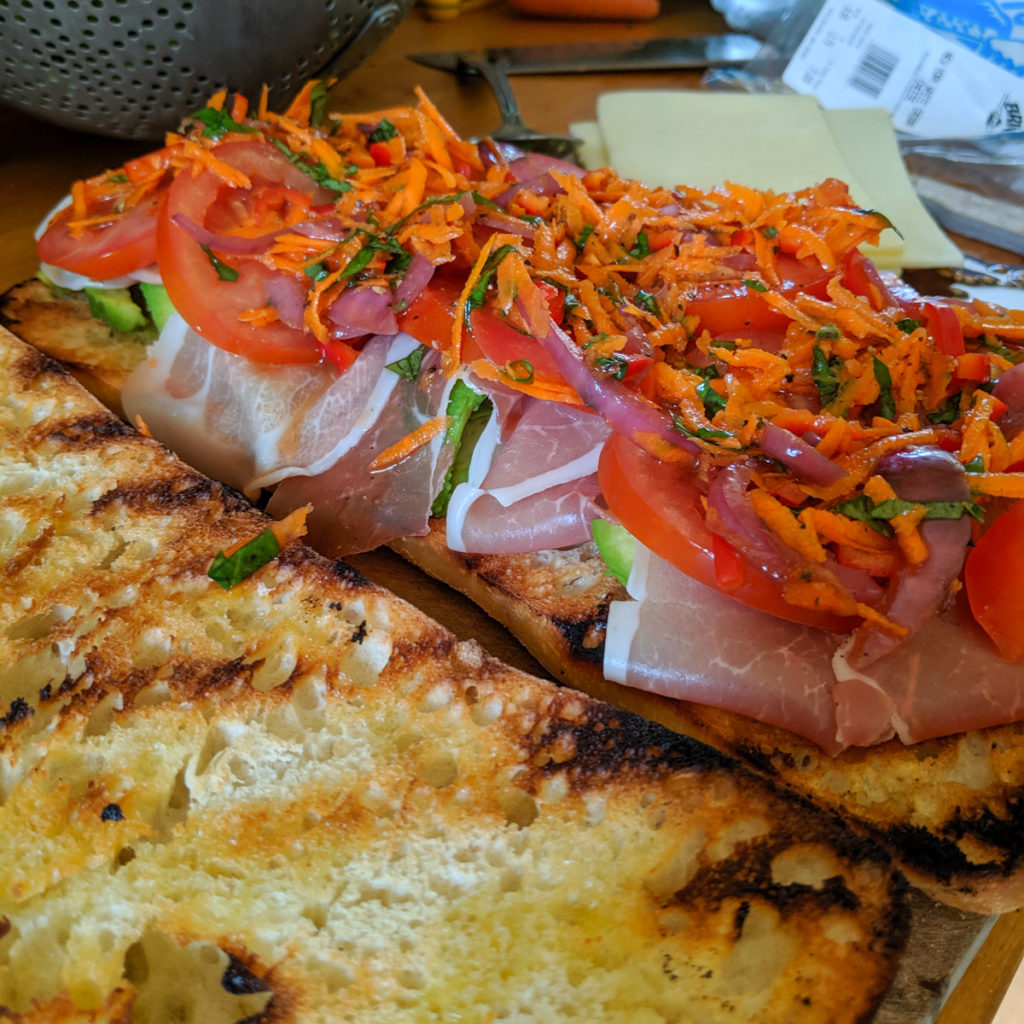 Sub sandwich on ciabatta being assembled with avocado, meat and colorful giardiniera.