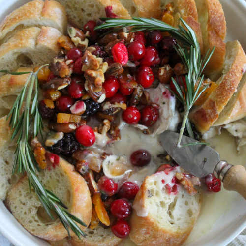 Cranberry Honey Baked Brie Appetizer surrounded by sliced baguette and garnished with rosemary.