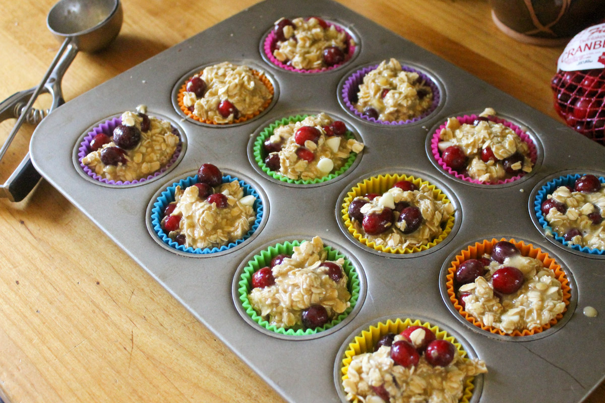 A muffin tin with colored liners full of cranberry batter with a portion scoop on the counter.