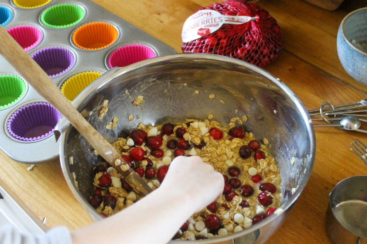 A child's hand mixing a bowl of cranberry oatmeal cup batter next to a muffin tin with colorful liners.