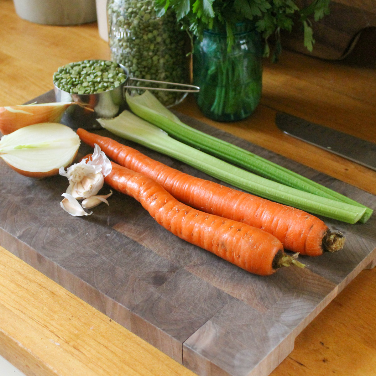 Split pea soup ingredients on a cutting board including raw carrots, celery, onion, garlic, and a jar with a measuring cup of dry split peas.