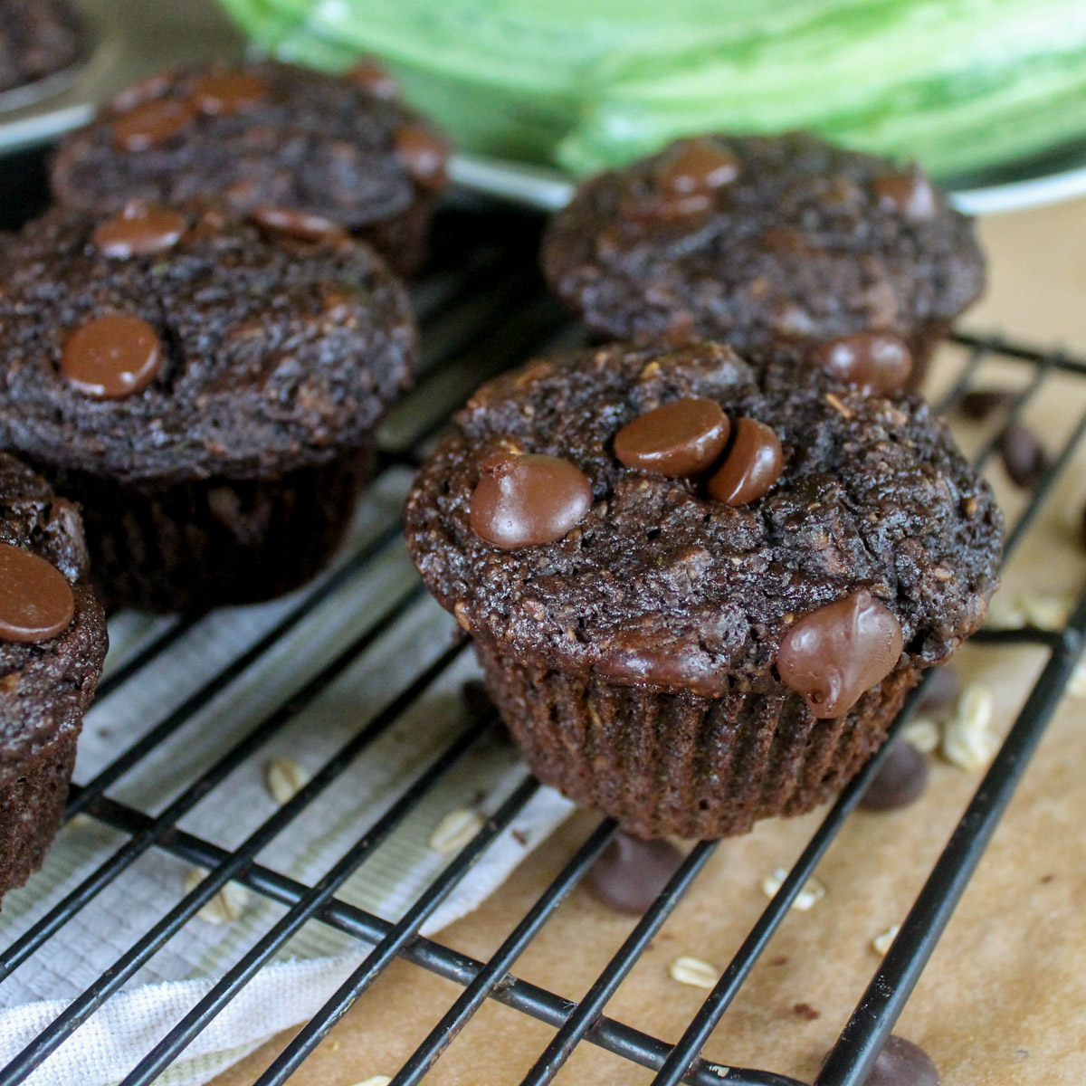 Chocolate zucchini oatmeal muffins in front of a bowl of green zucchinis.