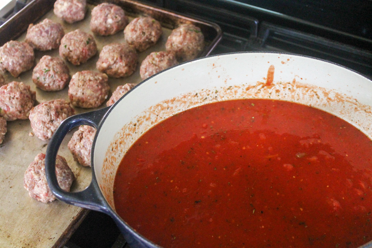 A sheet pan of meatballs ready to bake and a pot of marinara sauce ready to simmer on the stovetop.