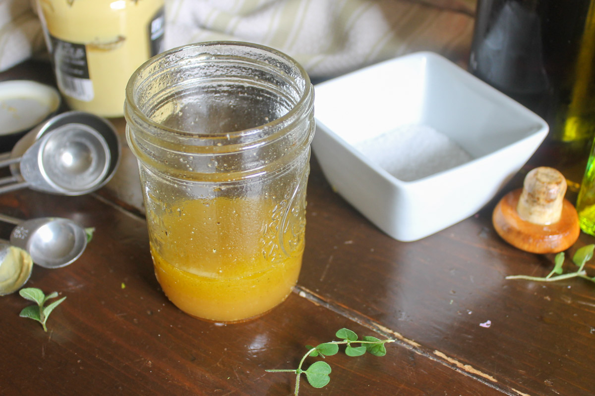 Salad dressing made in a jar in front of a bowl of salt, dijon mustard and measuring spoons.