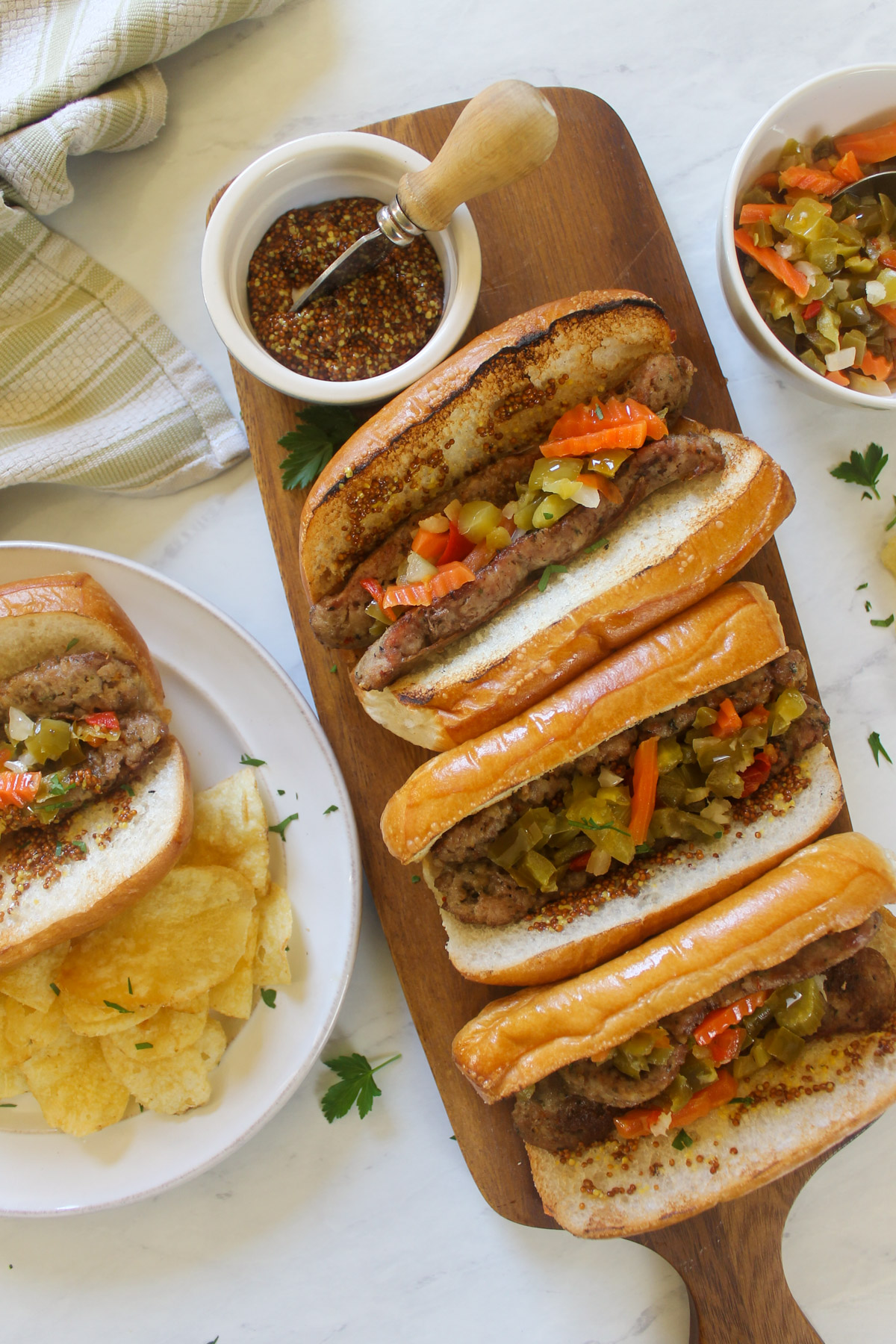 A wooden platter with 3 grilled Italian sausages in buns topped with giardiniera and a small bowl of grainy dijon mustard.