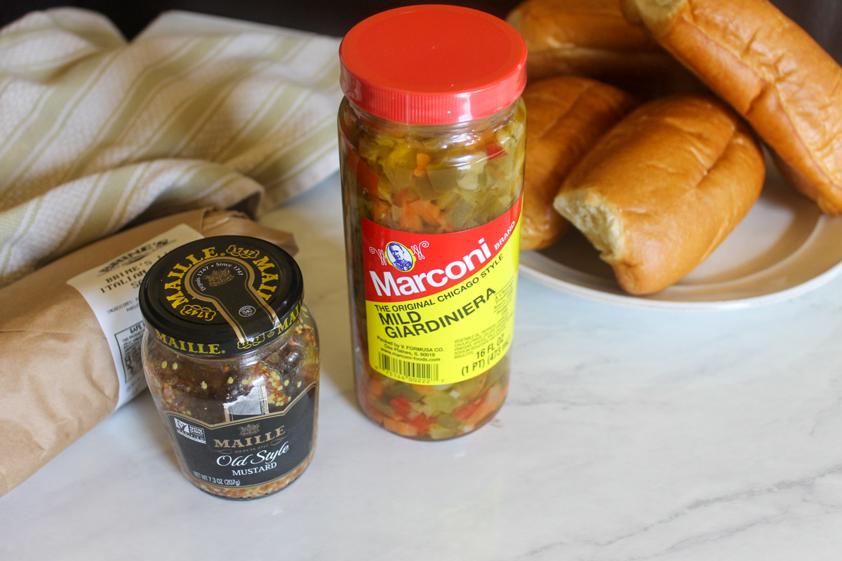 A jar of Dijon mustard and a jar of Giardiniera in front of a package of sausages and hoagie buns.