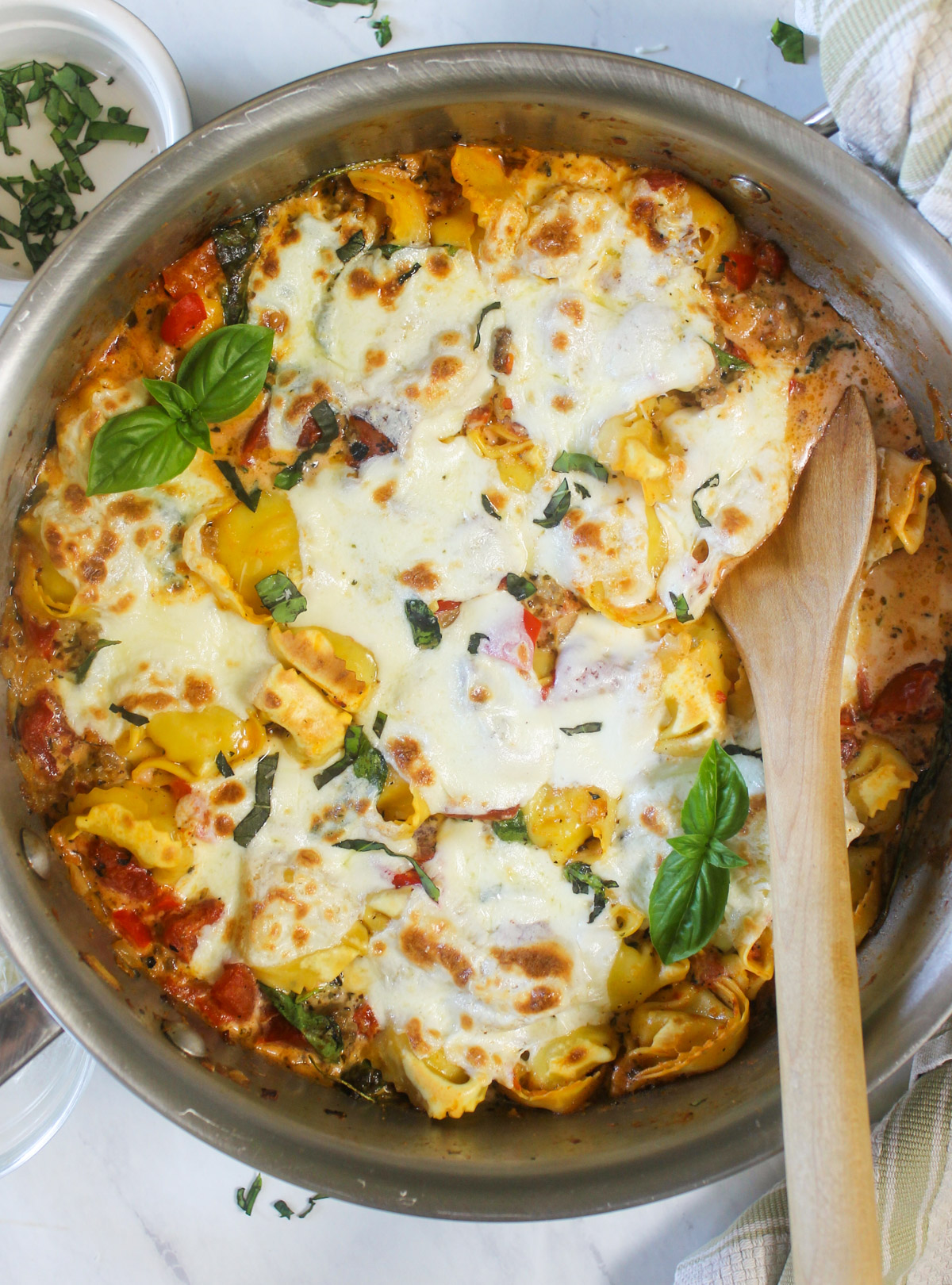 A finished pot of Baked Tortellini with Italian Sausage with a wooden spoon and basil leaves.