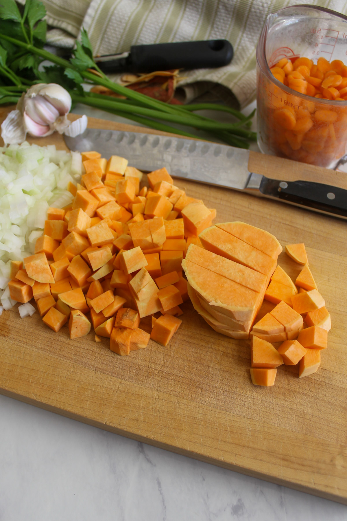 Chopped sweet potato, and onion on a cutting board with carrot, celery and garlic.
