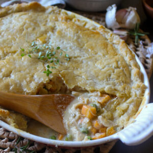 A white baking dish of chicken veggie pot pie with a flakey crust and a wooden spoon breaking into the filling.