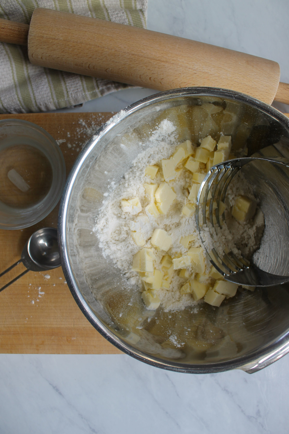 A bowl of flour and butter for pie crust on a cutting board with a rolling pin and bowl of water.